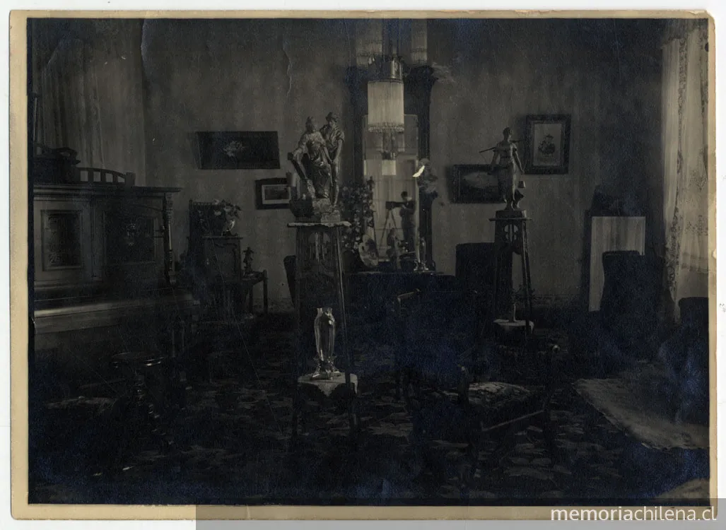 Salón con un piano, cuadros, pequeñas esculturas sobre peana, finos muebles, al fondo en un espejo se refleja la imagen del fotografo y su cámara — 1915 | Chile de Ayer