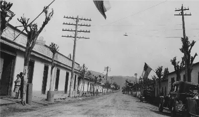 Calle principal de pueblo chileno engalanada con banderas y automóvil antiguo patente 16139 — Década de 1940 | Chile de Ayer