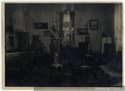 Salón con un piano, cuadros, pequeñas esculturas sobre peana, finos muebles, al fondo en un espejo se refleja la imagen del fotografo y su cámara — 1915 | Chile de Ayer