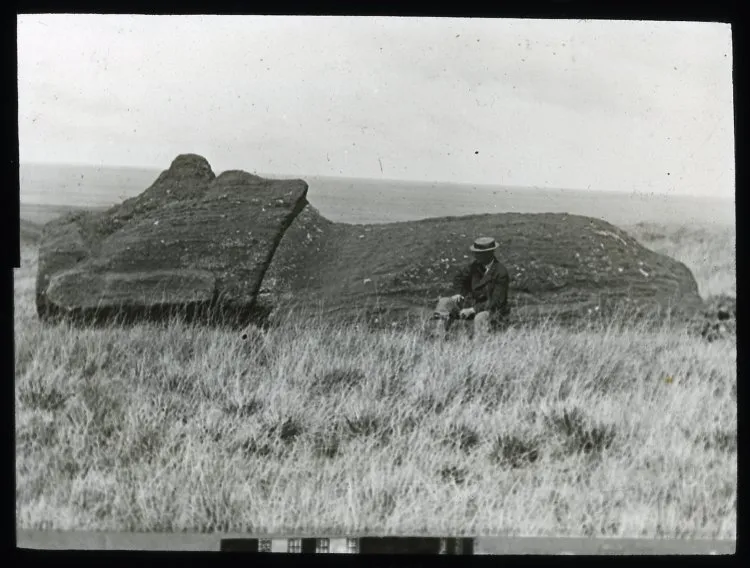 A moai lying on the ground, face upwards; on the moai statue road(?), with a White man wearing a hat and a moustache, sitting in front of it, for scale; Oc,G.T.1762, Mana Expedition to Easter Island, British Museum — Isla de Pascua — 1914 | Chile de Ayer