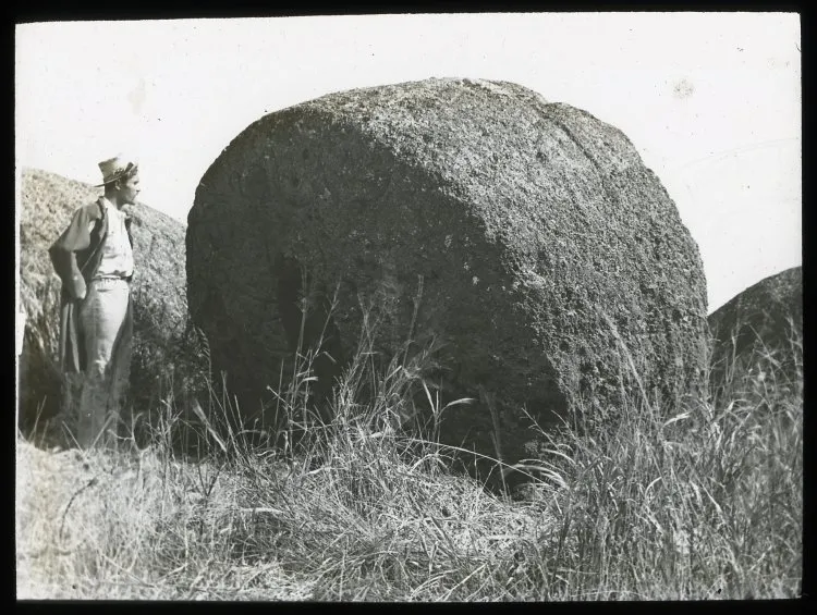 View of a large round stone, to be used as part of a moai, with a Polynesian man standing beside it, for scale; outside of Puna Pau, Oc,G.T.1626, Mana Expedition to Easter Island, British Museum — Isla de Pascua — 1914 | Chile de Ayer