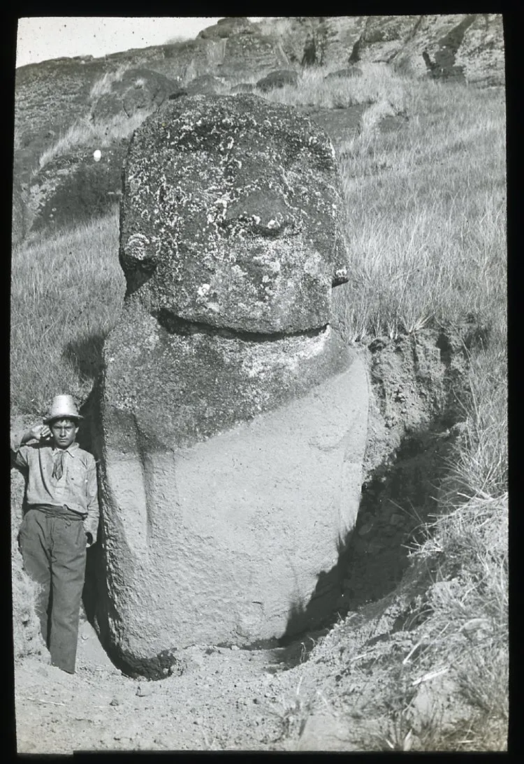 View of a moai, with its face eroded, partially excavated, standing upright in the ground, with a South American young man standing beside it on the left, for scale; Easter Island; Oc,G.T.1759, Mana Expedition to Easter Island, British Museum — Isla de Pascua — 1914 | Chile de Ayer