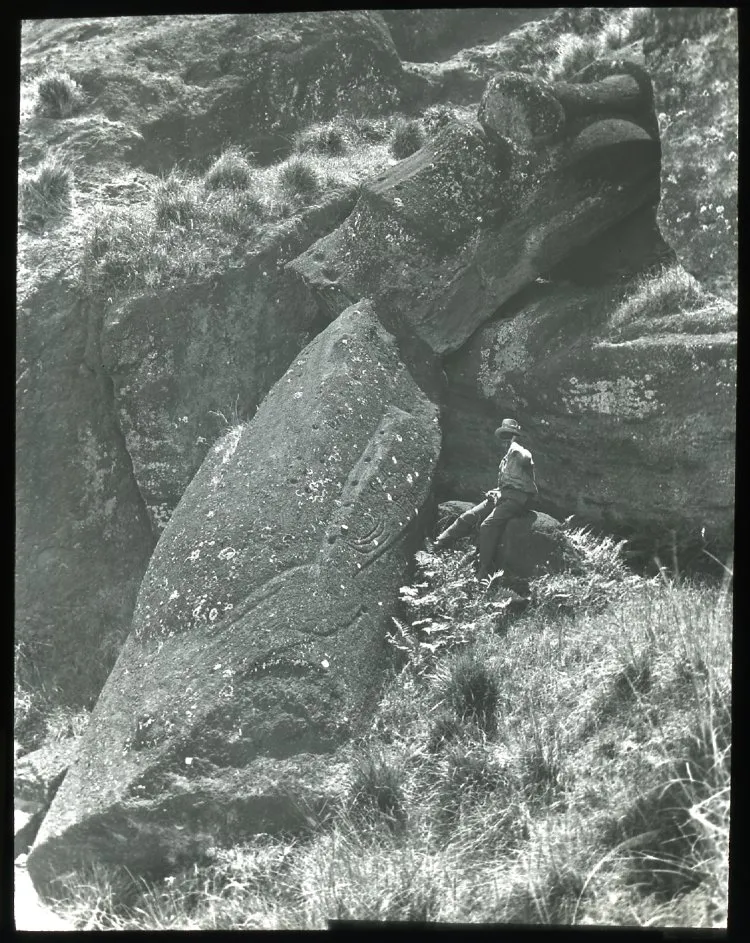 View of a moai having fallen and broken in two, on a steep slope with large rocks; a Chilean(?) boy sits beside the sculpture on the right, for scale; Oc,G.T.1763, Mana Expedition to Easter Island, British Museum — Isla de Pascua — 1914 | Chile de Ayer