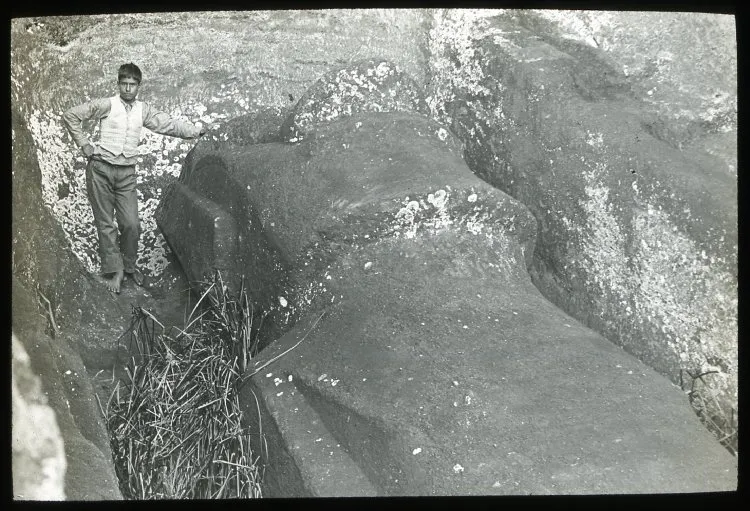 View of a moai lying in the ground with the boy standing on the left of the sculpture, resting one hand on the stone head, is for scale; Rano Raraku; Oc,G.T.1475, Mana Expedition to Easter Island, British Museum — Isla de Pascua — 1914 | Chile de Ayer