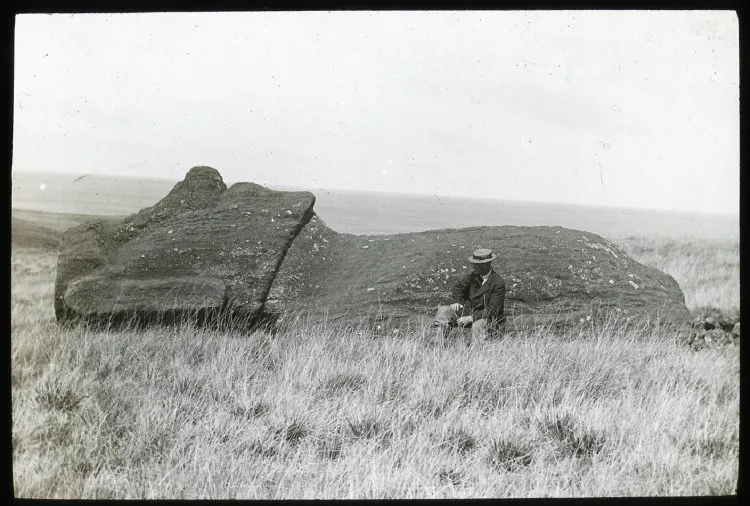 View of a moai lying on its back on the ground, with William Scoresby Routledge(?) sitting in front of the sculpture as a scale; Easter Island; Oc,G.T.1477, Mana Expedition to Easter Island, British Museum — Isla de Pascua — 1914 | Chile de Ayer