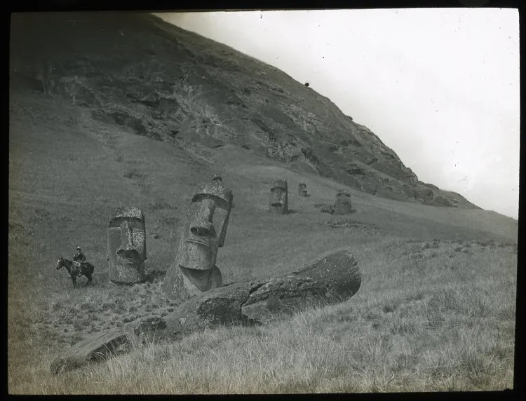 View of a moai lying on the ground, with five moai heads standing upright, buried in the ground; a man is sitting on a horse on the left of the image for scale; Rano Raraku, Oc,G.T.1478, Mana Expedition to Easter Island, British Museum — Isla de Pascua — 1914 | Chile de Ayer
