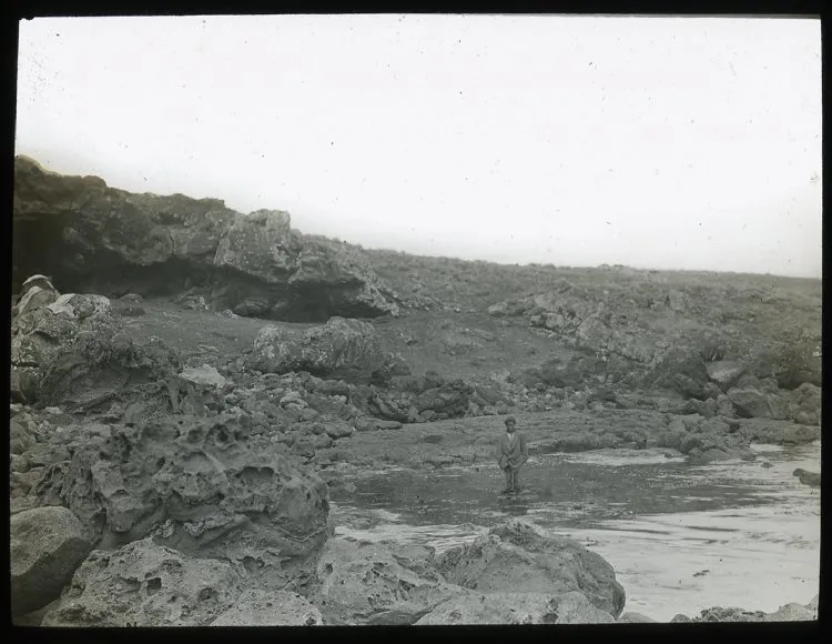 View of rocky shoreline, with entrance to Ana Havea, cave, on the left, and a Polyneisan(?) boy standing at the point where fresh water comes out, in the centre of the image; Oc,G.T.1630, Mana Expedition to Easter Island, British Museum — Isla de Pascua — 1914 | Chile de Ayer