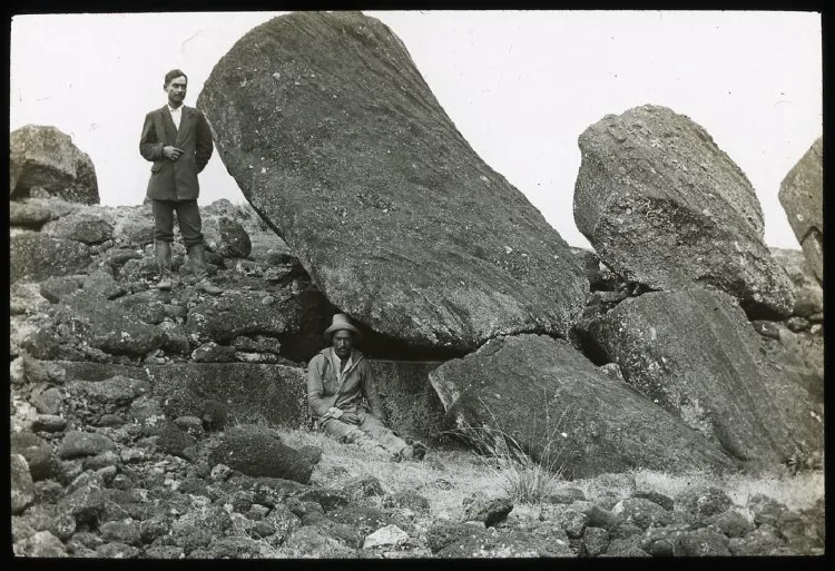 View of the back of two moai heads partially collapsed, with a man standing on the left of the first sculpture and a second man sitting beside the same sculpture for scale; Ahu Tongariki, Oc,G.T.1490, Mana Expedition to Easter Island, British Museum — Isla de Pascua — 1914 | Chile de Ayer