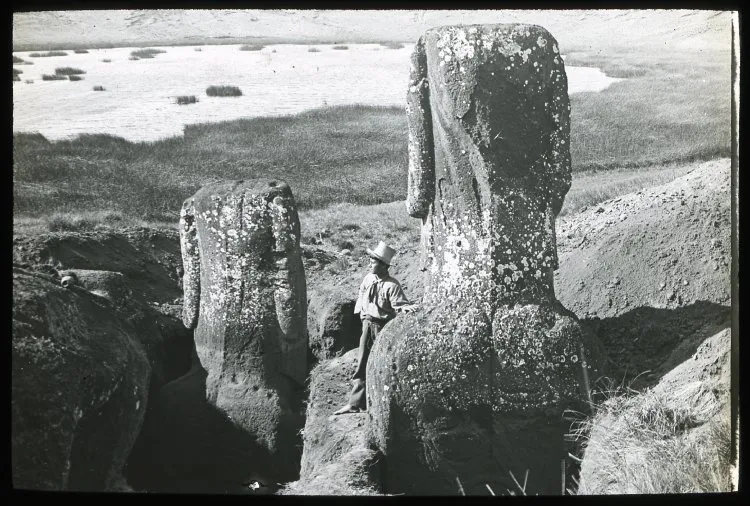 View of the back of two partially excavated moai with a Polynesian(?) man standing beside the figure in the foreground for scale; a lake in the background; Rano Raraku, Oc,G.T.1609, Mana Expedition to Easter Island, British Museum — Isla de Pascua — 1914 | Chile de Ayer