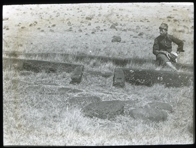 View of the foundation for the door and front of a hare paenga, with a Chilean(?) boy sitting on one of the stones, for scale; Easter Island, Oc,G.T.1638, Mana Expedition to Easter Island, British Museum — Isla de Pascua — 1914 | Chile de Ayer