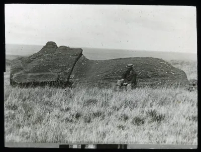 A moai lying on the ground, face upwards; on the moai statue road(?), with a White man wearing a hat and a moustache, sitting in front of it, for scale; Oc,G.T.1762, Mana Expedition to Easter Island, British Museum — Isla de Pascua — 1914 | Chile de Ayer