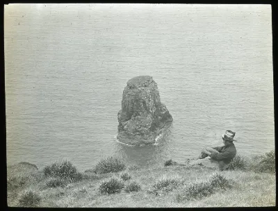 Chilean boy sitting down by cliff with view of Marotiri; Poike, Oc,G.T.1627, Mana Expedition to Easter Island, British Museum — Isla de Pascua — 1914 | Chile de Ayer