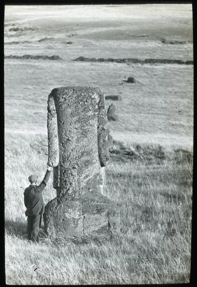 View of a back of moai with a man standing on the left of the sculpture with his back to the camera and one hand on the left earlobe, for scale; Rano Raraku; Oc,G.T.1474, Mana Expedition to Easter Island, British Museum — Isla de Pascua — 1914 | Chile de Ayer