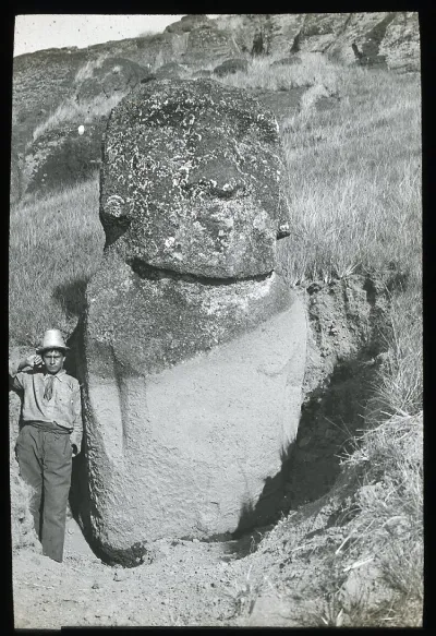 View of a moai, with its face eroded, partially excavated, standing upright in the ground, with a South American young man standing beside it on the left, for scale; Easter Island; Oc,G.T.1759, Mana Expedition to Easter Island, British Museum — Isla de Pascua — 1914 | Chile de Ayer