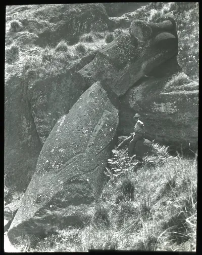 View of a moai having fallen and broken in two, on a steep slope with large rocks; a Chilean(?) boy sits beside the sculpture on the right, for scale; Oc,G.T.1763, Mana Expedition to Easter Island, British Museum — Isla de Pascua — 1914 | Chile de Ayer
