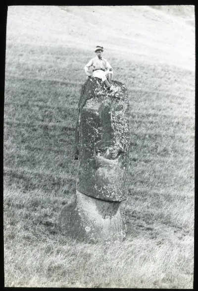 View of a moai head standing upright in the ground, with a boy sitting on top of the carved head for scale; Rano Raraku, Oc,G.T.1467, Mana Expedition to Easter Island, British Museum — Isla de Pascua — 1914 | Chile de Ayer