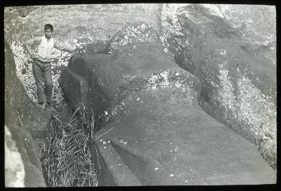 View of a moai lying in the ground with the boy standing on the left of the sculpture, resting one hand on the stone head, is for scale; Rano Raraku; Oc,G.T.1475, Mana Expedition to Easter Island, British Museum — Isla de Pascua — 1914 | Chile de Ayer