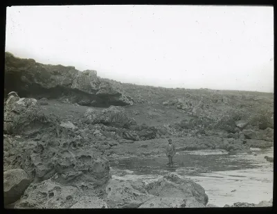 View of rocky shoreline, with entrance to Ana Havea, cave, on the left, and a Polyneisan(?) boy standing at the point where fresh water comes out, in the centre of the image; Oc,G.T.1630, Mana Expedition to Easter Island, British Museum — Isla de Pascua — 1914 | Chile de Ayer