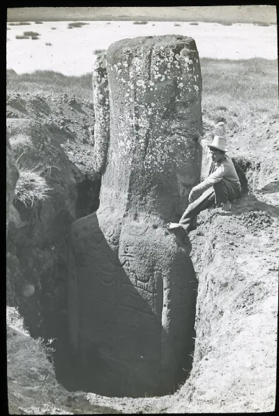 View of the back of a moai with petroglyph carvings, partially excavated; a South American man is sitting on the ground, with one bare foot on the shoulder of the sculpture; Rano Raraku, Oc,G.T.1465, Mana Expedition to Easter Island, British Museum — Isla de Pascua — 1914 | Chile de Ayer