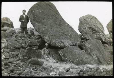 View of the back of two moai heads partially collapsed, with a man standing on the left of the first sculpture and a second man sitting beside the same sculpture for scale; Ahu Tongariki, Oc,G.T.1490, Mana Expedition to Easter Island, British Museum — Isla de Pascua — 1914 | Chile de Ayer