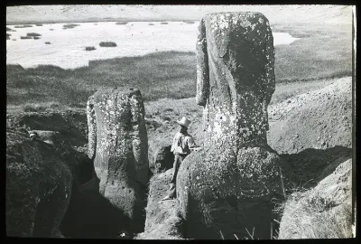 View of the back of two partially excavated moai with a Polynesian(?) man standing beside the figure in the foreground for scale; a lake in the background; Rano Raraku, Oc,G.T.1609, Mana Expedition to Easter Island, British Museum — Isla de Pascua — 1914 | Chile de Ayer