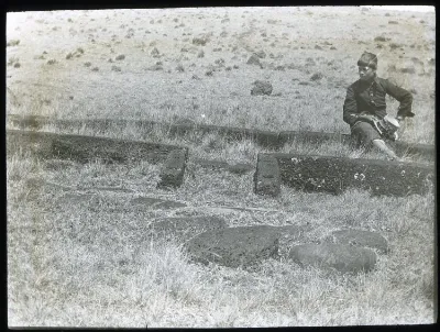 View of the foundation for the door and front of a hare paenga, with a Chilean(?) boy sitting on one of the stones, for scale; Easter Island, Oc,G.T.1638, Mana Expedition to Easter Island, British Museum — Isla de Pascua — 1914 | Chile de Ayer
