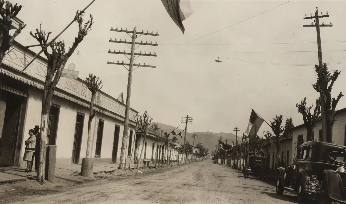 Calle principal de pueblo chileno engalanada con banderas y automóvil antiguo patente 16139 — Década de 1940 | Chile de Ayer