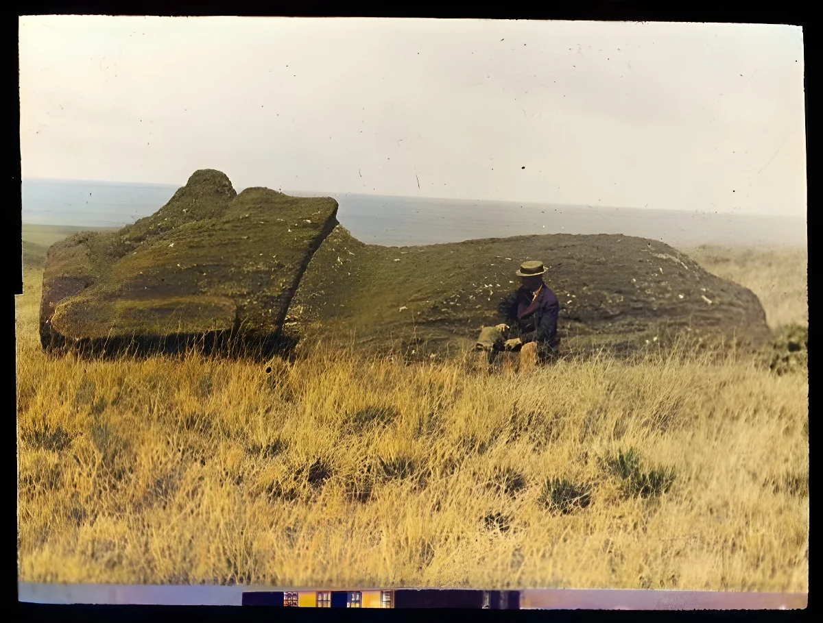 A moai lying on the ground, face upwards; on the moai statue road(?), with a White man wearing a hat and a moustache, sitting in front of it, for scale; Oc,G.T.1762, Mana Expedition to Easter Island, British Museum — Isla de Pascua — 1914 | Chile de Ayer