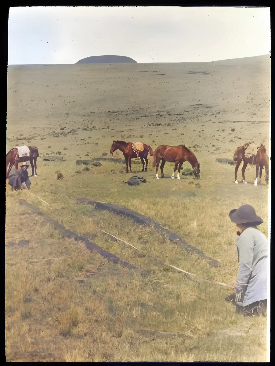 Two Polynesian(?) men kneeling on the ground, measuring the length of the foundations of a hare paenga; four horses grazing in the background; slopes of Poike visible in background; Oc,G.T.1637, Mana Expedition to Easter Island, British Museum — Isla de Pascua — 1914 | Chile de Ayer