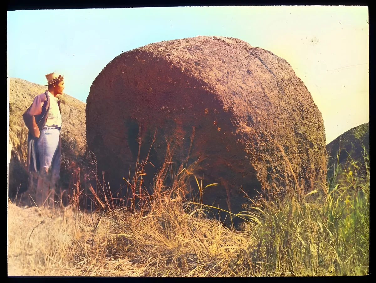 View of a large round stone, to be used as part of a moai, with a Polynesian man standing beside it, for scale; outside of Puna Pau, Oc,G.T.1626, Mana Expedition to Easter Island, British Museum — Isla de Pascua — 1914 | Chile de Ayer