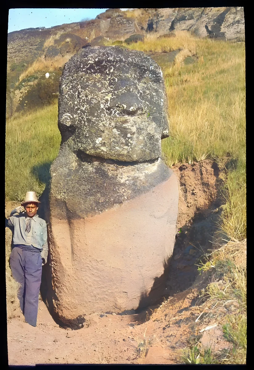View of a moai, with its face eroded, partially excavated, standing upright in the ground, with a South American young man standing beside it on the left, for scale; Easter Island; Oc,G.T.1759, Mana Expedition to Easter Island, British Museum — Isla de Pascua — 1914 | Chile de Ayer