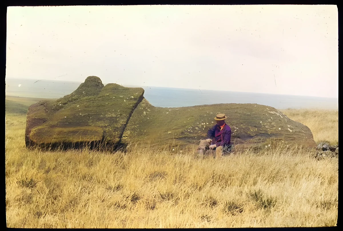 View of a moai lying on its back on the ground, with William Scoresby Routledge(?) sitting in front of the sculpture as a scale; Easter Island; Oc,G.T.1477, Mana Expedition to Easter Island, British Museum — Isla de Pascua — 1914 | Chile de Ayer