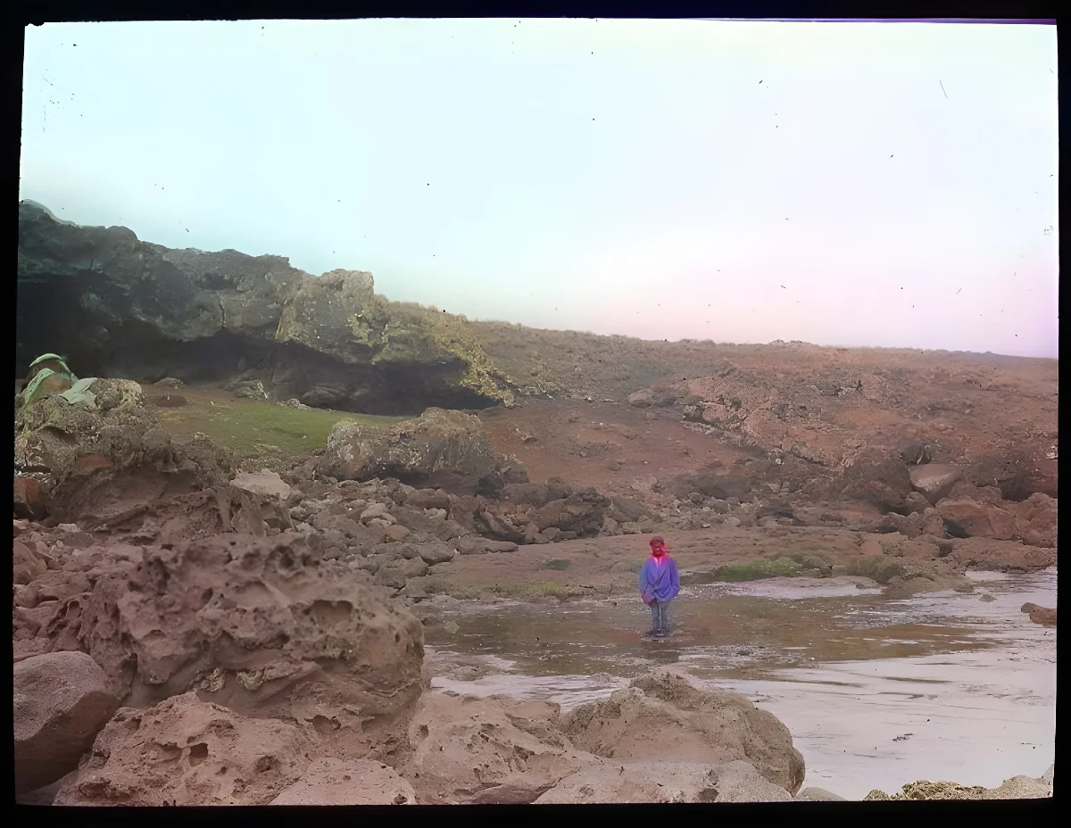 View of rocky shoreline, with entrance to Ana Havea, cave, on the left, and a Polyneisan(?) boy standing at the point where fresh water comes out, in the centre of the image; Oc,G.T.1630, Mana Expedition to Easter Island, British Museum — Isla de Pascua — 1914 | Chile de Ayer