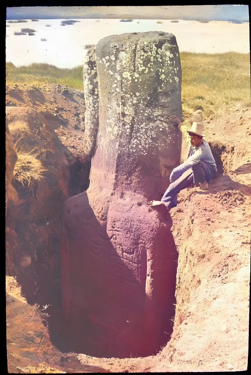 View of the back of a moai with petroglyph carvings, partially excavated; a South American man is sitting on the ground, with one bare foot on the shoulder of the sculpture; Rano Raraku, Oc,G.T.1465, Mana Expedition to Easter Island, British Museum — Isla de Pascua — 1914 | Chile de Ayer