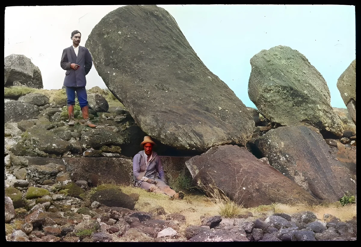 View of the back of two moai heads partially collapsed, with a man standing on the left of the first sculpture and a second man sitting beside the same sculpture for scale; Ahu Tongariki, Oc,G.T.1490, Mana Expedition to Easter Island, British Museum — Isla de Pascua — 1914 | Chile de Ayer