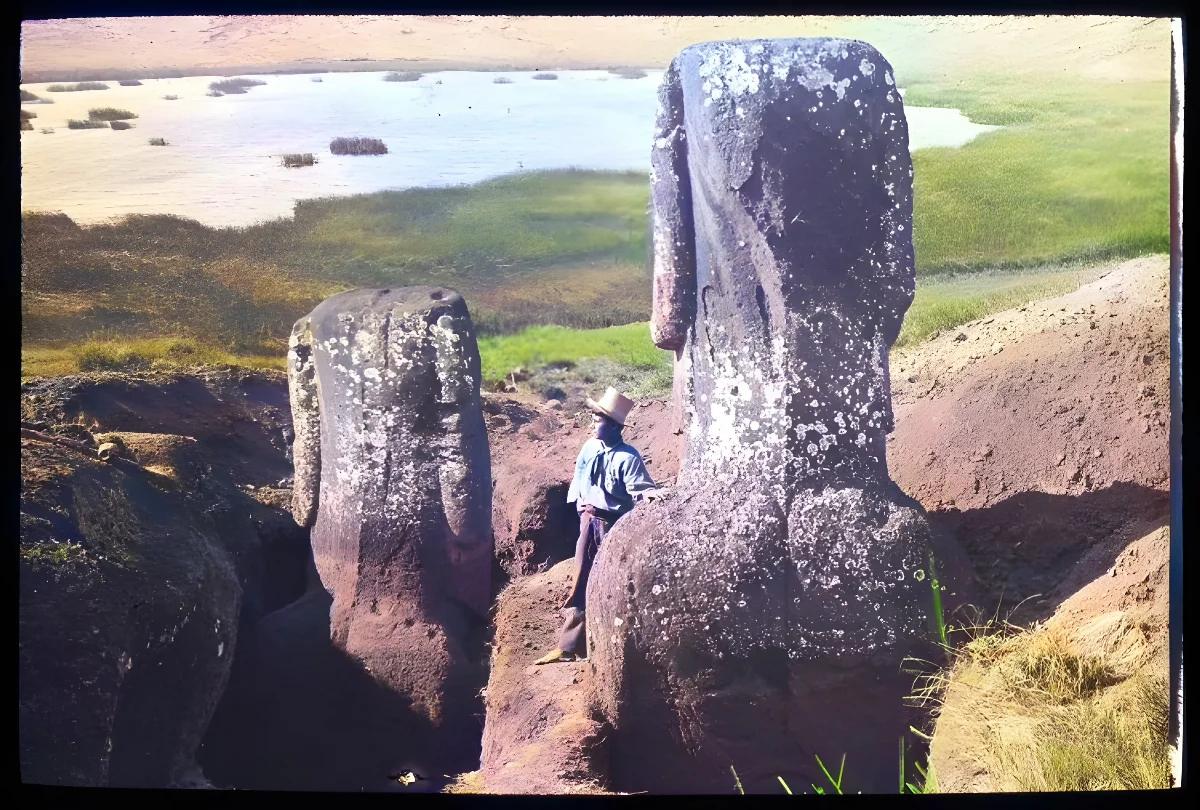 View of the back of two partially excavated moai with a Polynesian(?) man standing beside the figure in the foreground for scale; a lake in the background; Rano Raraku, Oc,G.T.1609, Mana Expedition to Easter Island, British Museum — Isla de Pascua — 1914 | Chile de Ayer