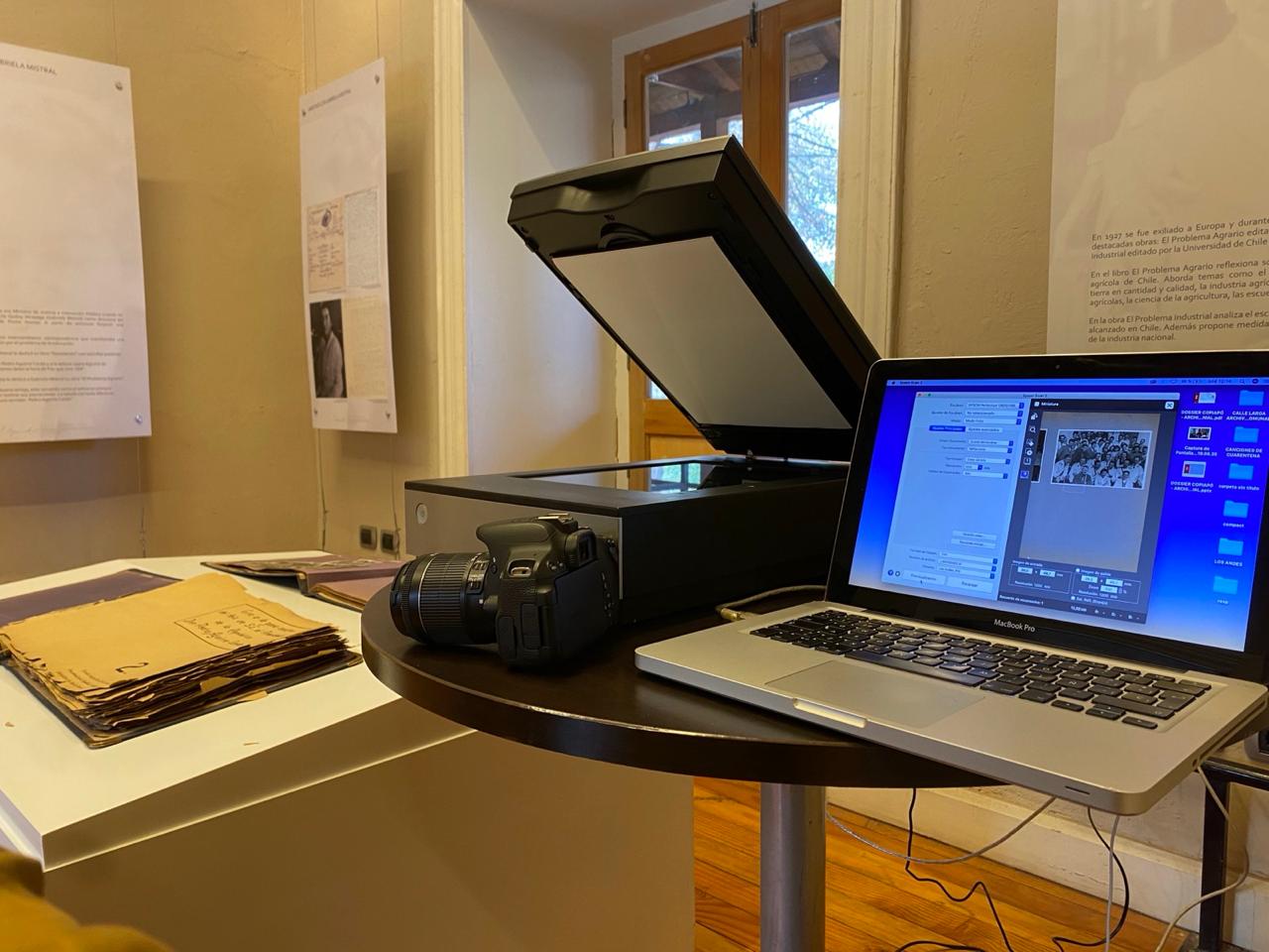 Setup de digitalización archivística: un escáner plano Epson Perfection V600, una cámara réflex, un laptop con software de captura y, en primer plano, una carpeta con documentos antiguos amarillentos. Al fondo, paneles de exposición sobre Gabriela Mistral.