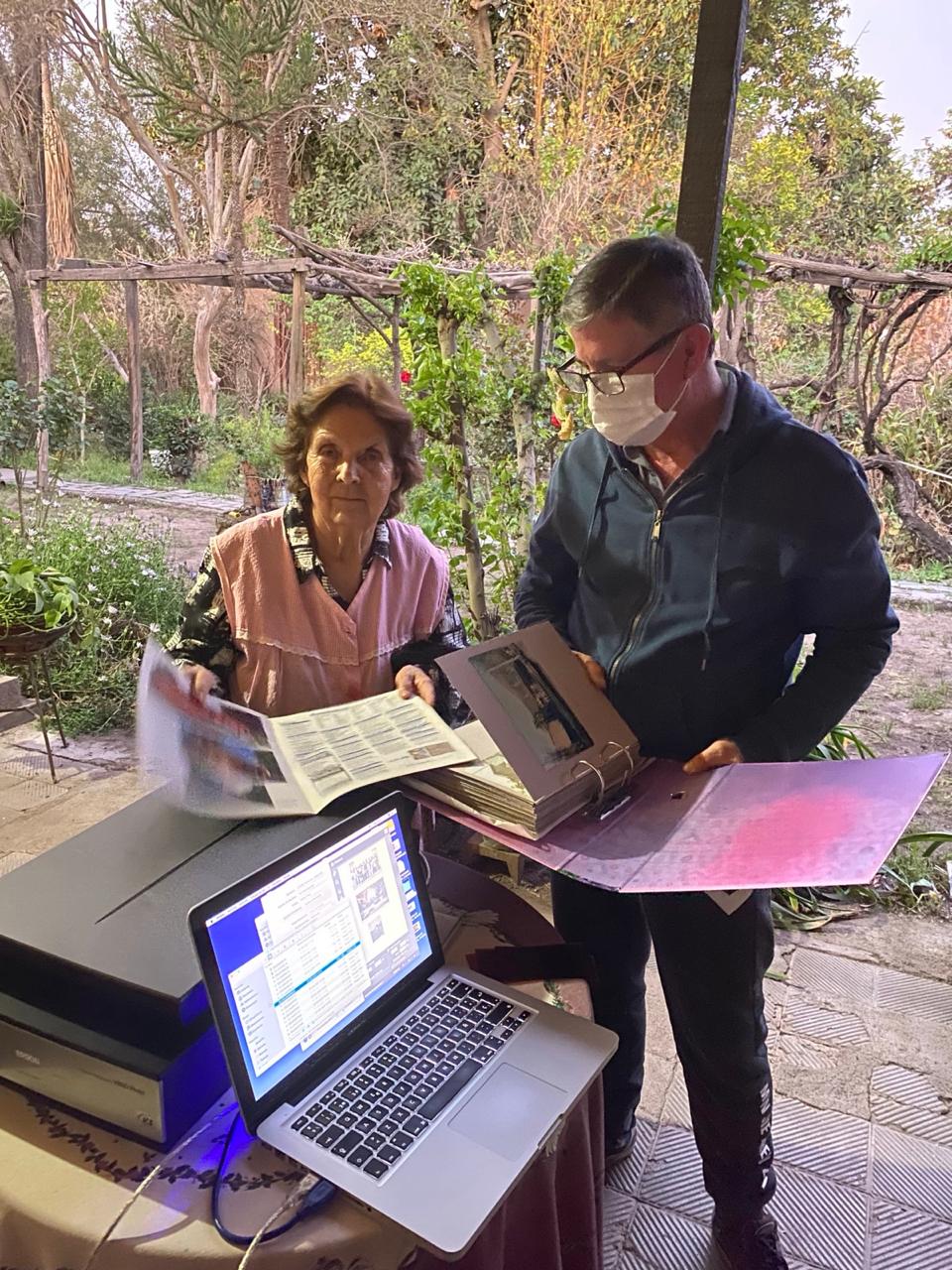 Una mujer mayor junto a un hombre con mascarilla y chaqueta oscura revisan un álbum abierto en un jardín con enredaderas. Sobre una mesa hay un escáner y un laptop encendido con el software de digitalización.