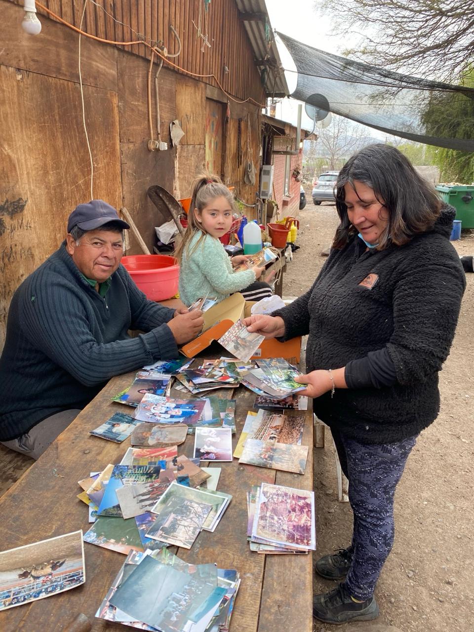 Una familia selecciona fotografías sobre una mesa de madera al aire libre. Un hombre con gorro, una niña y una mujer revisan decenas de fotos repartidas entre ellos.